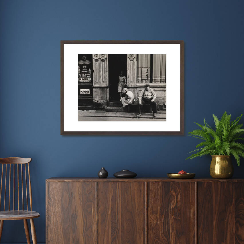 Zwart-witfoto uit omstreeks 1956 van twee meisjes en een man voor hun woonhuis in de Anjelierstraat in de Jordaan in Amsterdam. – donkerbruine wissellijst in woonkamer