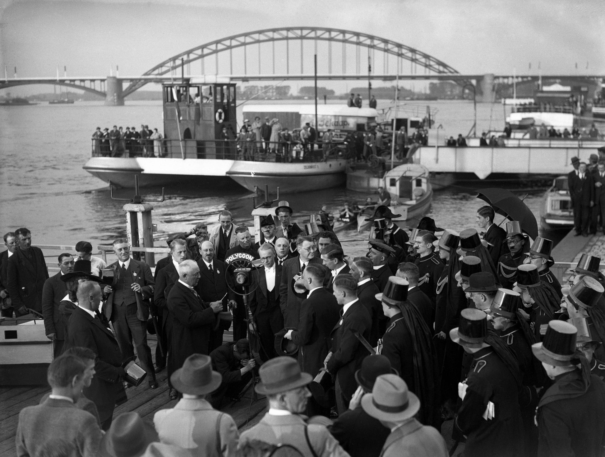 Zwart-witfoto van een carnavalsplechtigheid op de Waalkade in Nijmegen met de Waalbrug op de achtergrond.